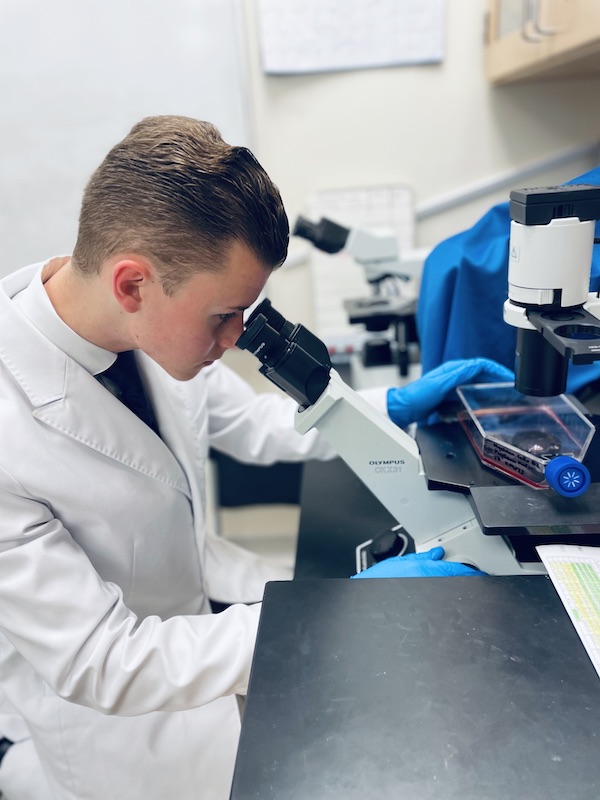 Riley in lab gear looking in a microscope in a laboratory.