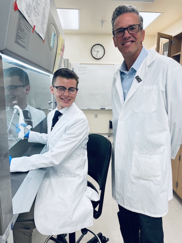 Gillis sits in a lab chair and Dr. AuCoin stands behind. Both smile at the camera while wearing lab gear.