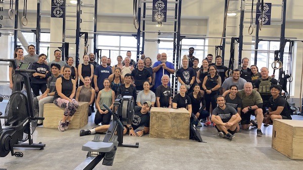 A large group of people in workout clothes smile for the camera in the functional training area of the fitness center.