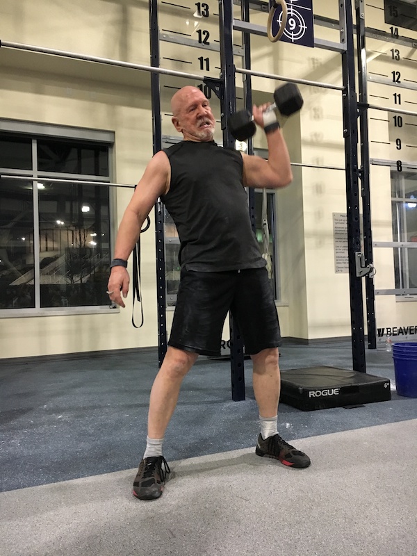A man holds a dumbell in one arm, brining it up past his shoulder, inside a fitness center.