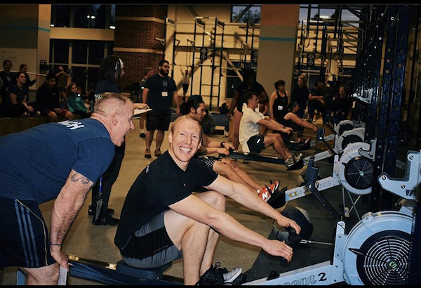 A man smiles broadly on the row machine while a trainer tells him something close to his face.