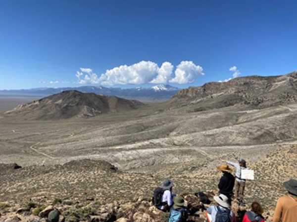 A group of researchers stand and gaze out at a massive, mountainous desert landscape.