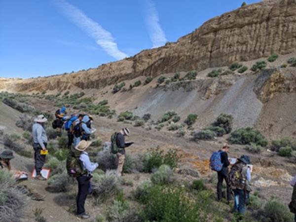 Researchers in hads with backbacks gaze upon a geographical cliff from the bottom looking up with various features present like gravel and water marks forming ridges. They are all taking notes in notebooks.