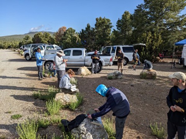 Students in field gear on a rugged terrain taking notes while sitting in dirt and on rocks.