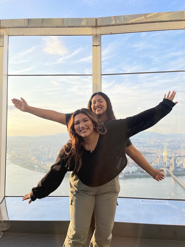 Lesly and Noemi pose one in front of the other with outstretched arms in front of a large window looking over a city.