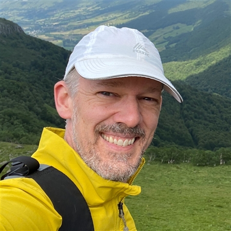 Tom Albright poses outside in rolling green hills smiling at the camera wearing a yellow sweater and a white baseball cap.