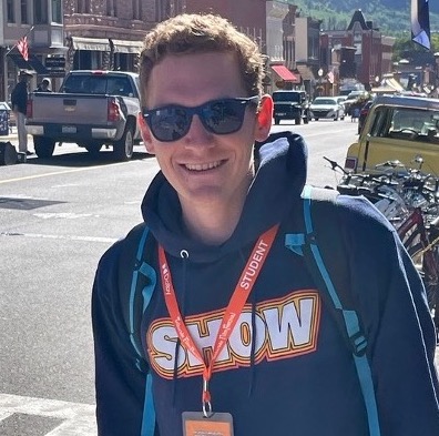 John Snelgrove in sunglasses and a sweatshirt smiles outdoors.