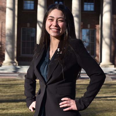 Lucy Burnham on the quad wearing a blazer with her hands on her hips, smiling.