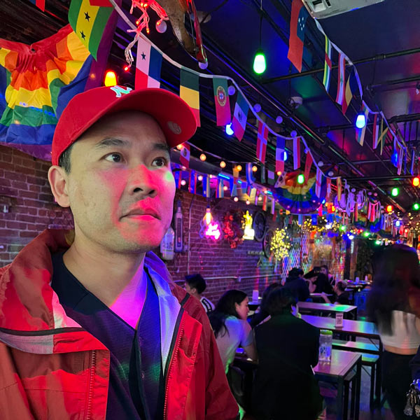 Paul Kwon on a night lit street with colorful banners hanging around. He's wearing a red cap and staring off into the distance.