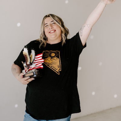Sadie Fienberg smiling in a black t-shirt with one hand raised in the air and the other holding an award with an eagle and American flag.