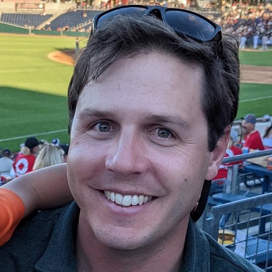 Andrew Zuza headshot smiling in a baseball stadium.
