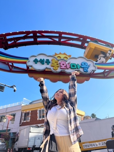 Lesly at a theme park with a Korean sign above her. She stands looking up, smiling, holding her hands up toward the sign.