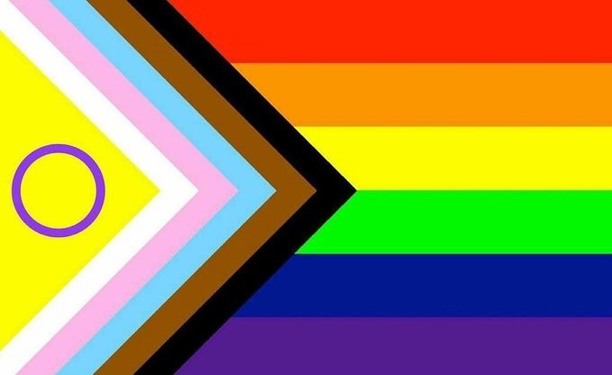 The Progress flag with added purple circle on a yellow field on the left side of the flag, set within white, pink, light blue, brown, and black arrows, which are pointing to the right, where the traditional rainbow Pride flag horizontal stripes of red, orange, yellow, green, blue, and purple sit.