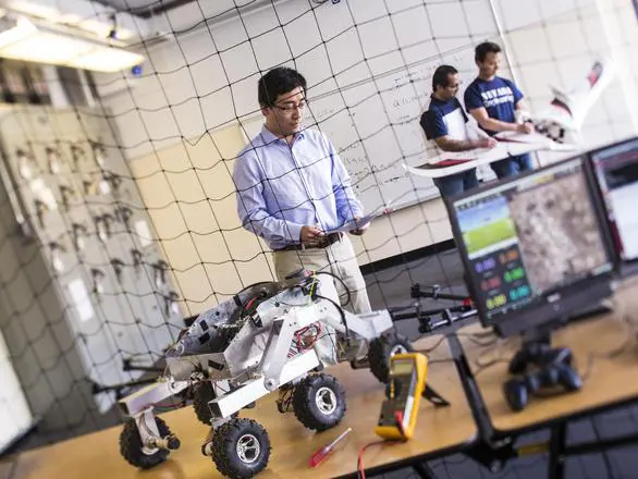 Three researchers work in the background of a drone sitting on a desk next to a desktop computer.