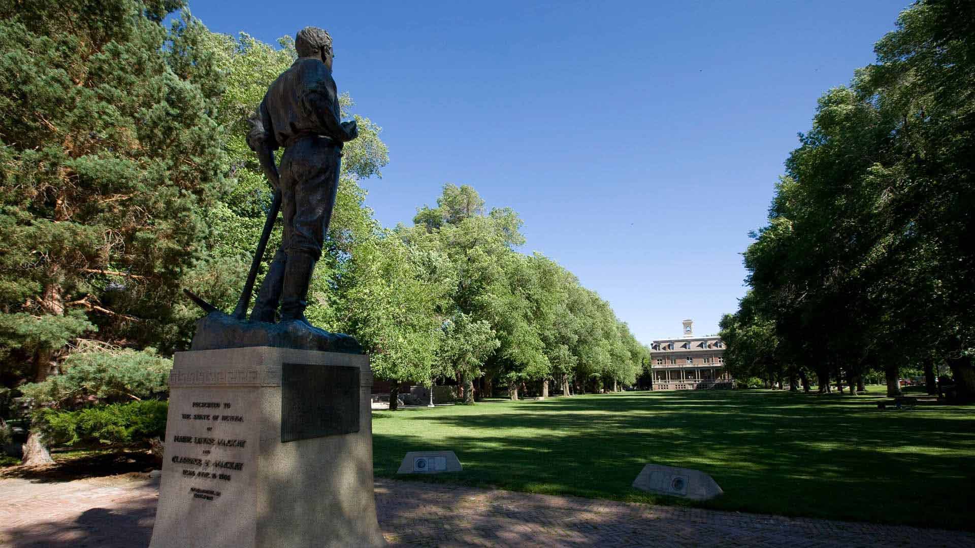 The John Mackay statue stands at the north end of the University of Nevada, Reno Quad -- a large lawn area surrounded by trees and an outer ring of buildings -- with Morrill Hall visible at the opposite end of the statue.