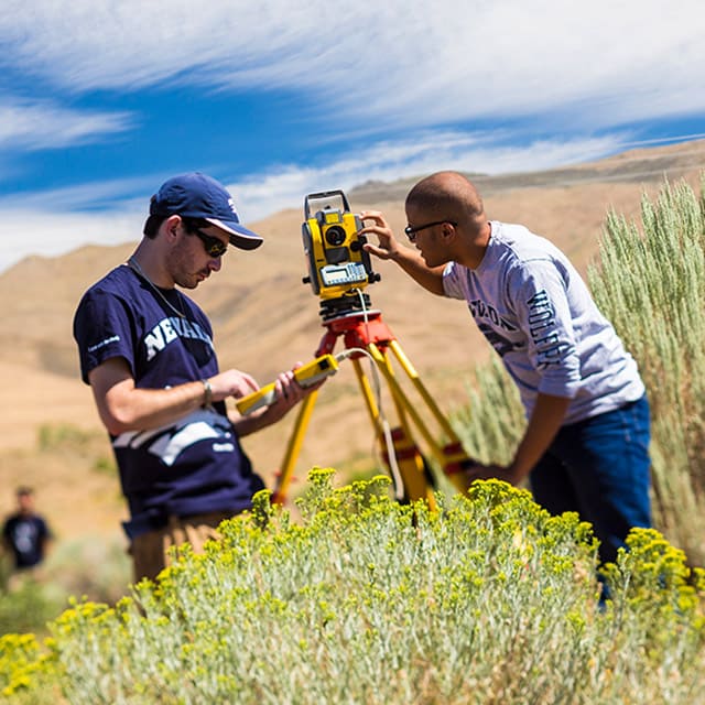Two people look through surveying equipment in a field.