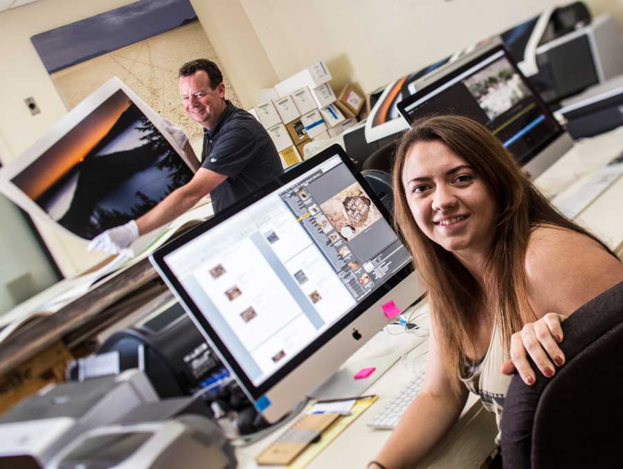 An art student in front of a computer with photo editiing software; in the background an instructor holds up a large print of Lake Tahoe at sunset