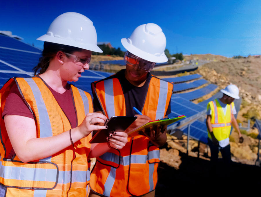 Two students in hard hats and reflective vests consult iPads and a clipboard at a field of solar panels