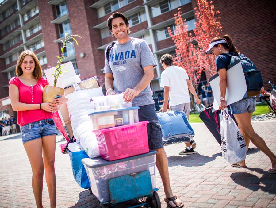 A student holding a potted plant and a parent stand outside a dorm, next to a loaded moving dolly