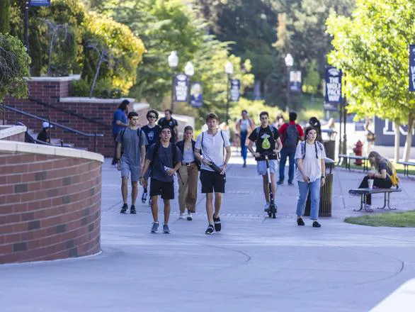 Several students walk with backpacks, while another rides a scooter, next to the Mathewson-IGT Knowledge Center on the University of Nevada, Reno campus.