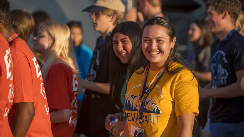 A student wearing a yellow "NevadaFIT" shirt and lanyard smiles for a picture while other NevadaFIT attendees walk around her.