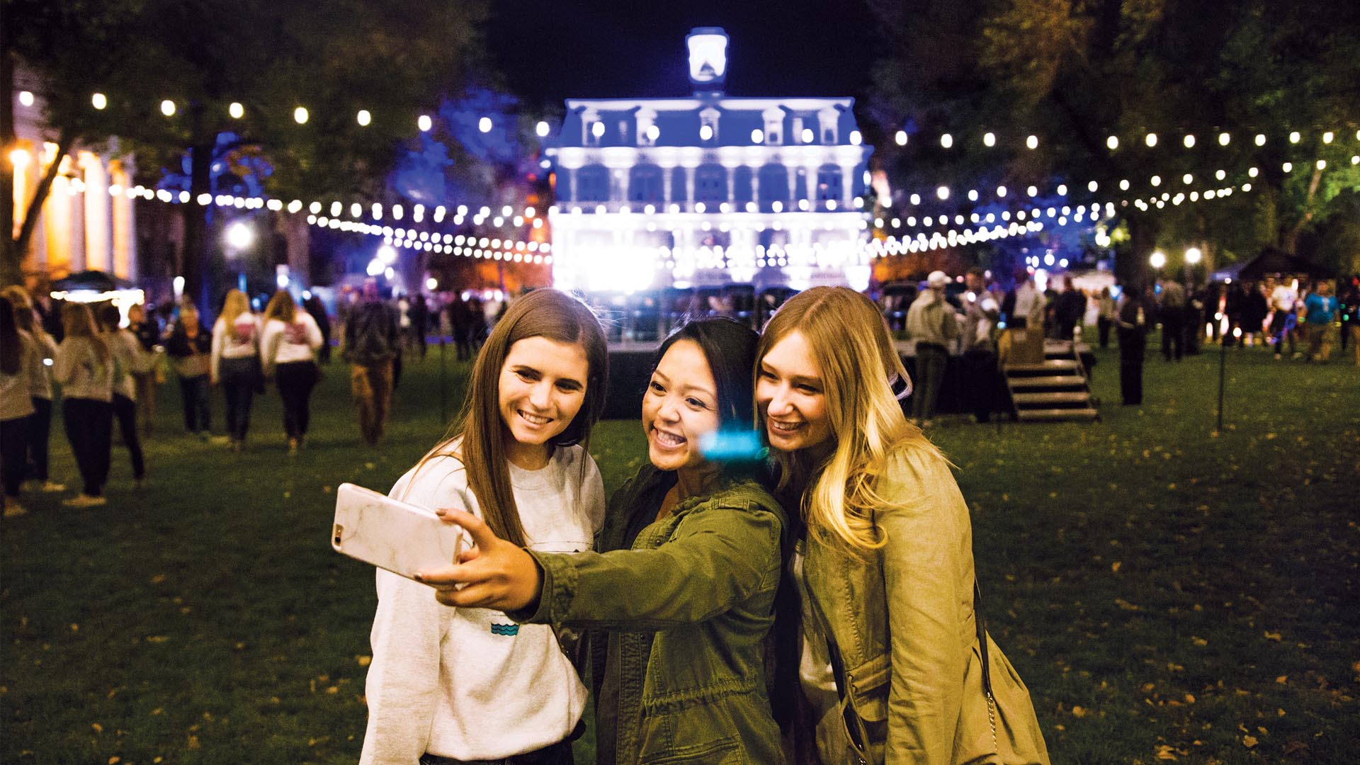 Three students pose for a selfie on the Quad at night in front of a lit-up Mackay Hall.