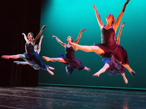 A group of four ballet dancers jump in the air and raise their arms during a performance on a stage lit with a green backdrop.