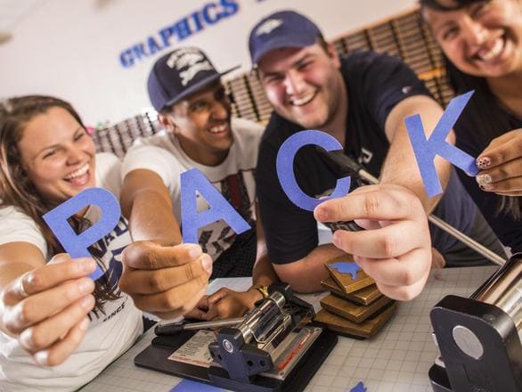 A group of four students smile and each individually hold up blue paper letters that spell the word Pack.