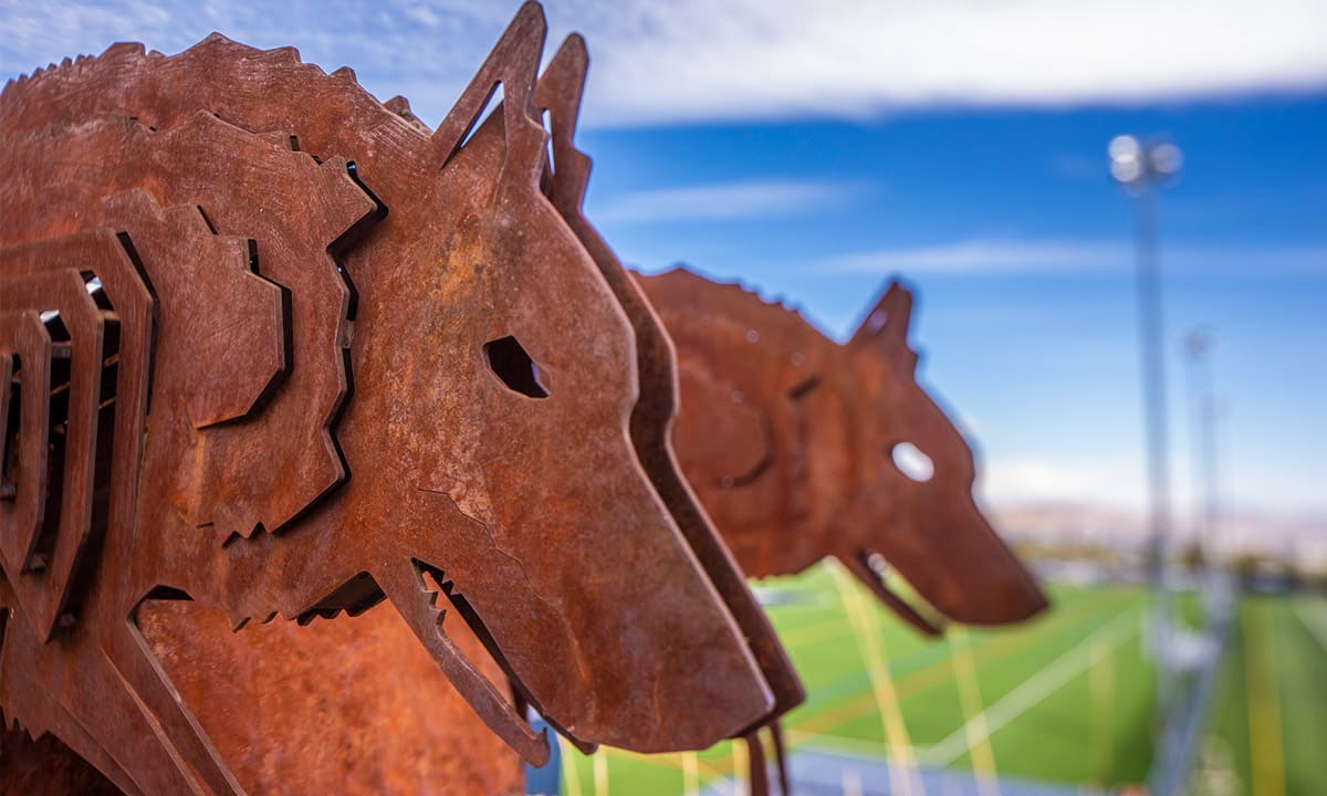 Close-up view of a rust-colored metal sculpture depicting stylized wolf heads, with sharp, angular features. The artwork is positioned outdoors overlooking a green sports field under a bright blue sky, symbolizing school spirit and the Wolf Pack identity at the University of Nevada, Reno.