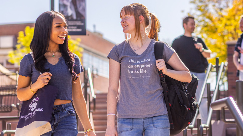 Two people standing outdoors in front of the Joe Crowley Student Union. The person on the left wears a blue crop top and jeans, accessorized with a necklace and a shoulder bag. The person on the right wears a navy blue sweatshirt with 'University of Nevada' printed on it. Behind them are trees and the JCSU lawn.
