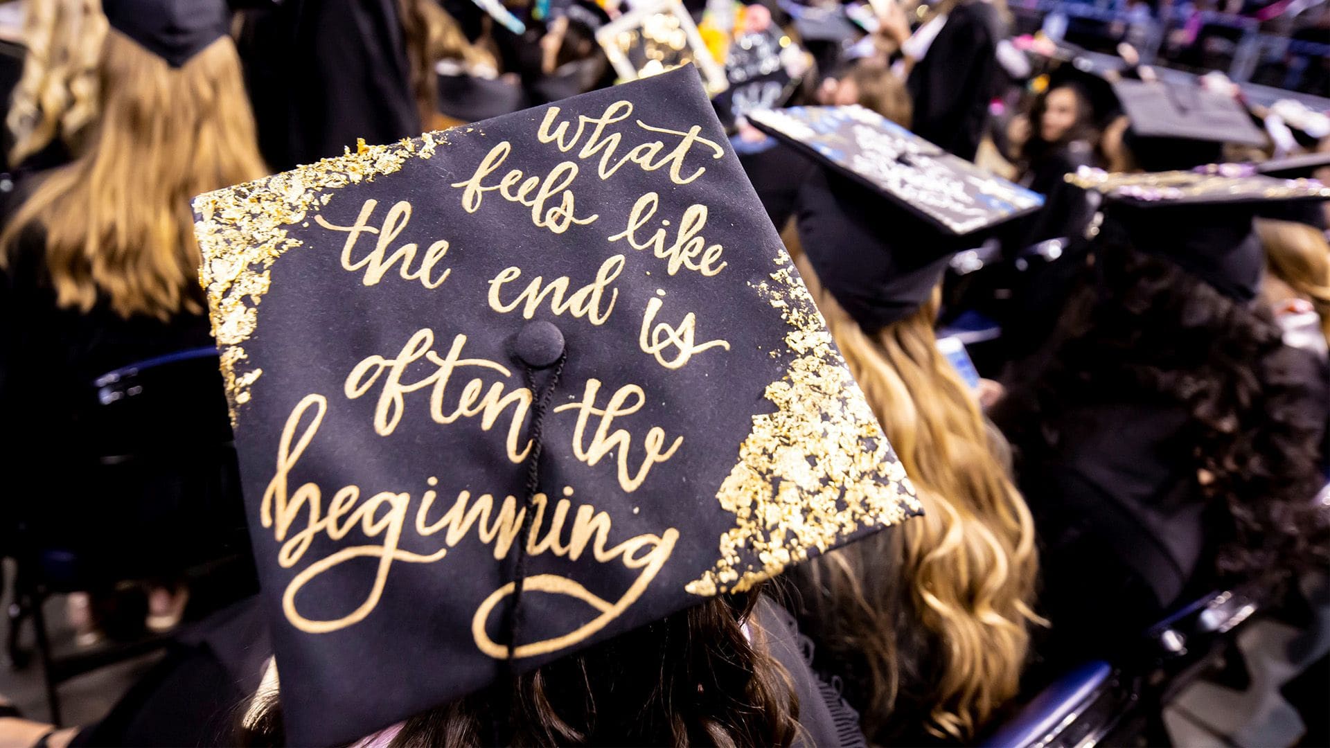 A student wearing a mortar board that reads "What feels like the end is often the beginning" sits next to other graduating students during a commencement ceremony.