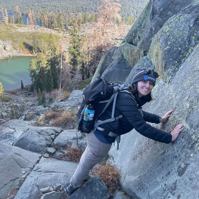 Gigi Bucciachio wears a hiking backpack and rests her arms against a giant boulder in the Tahoe wilderness.