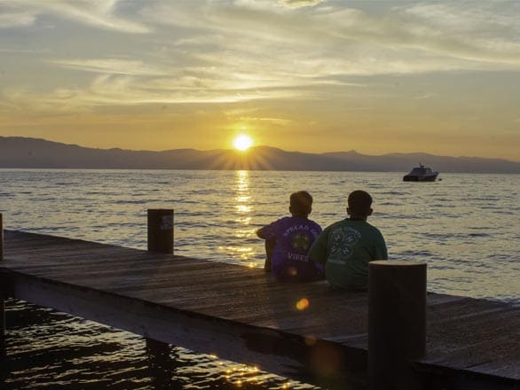 Two people seated on a wooden pier overlooking Lake Tahoe at sunset, with golden light reflecting on calm water and a boat in the distance.