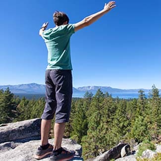 Person standing on a rocky outcrop overlooking Lake Tahoe with mountains in the distance.