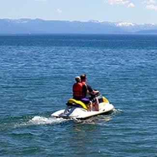 Two people riding a jet ski across Lake Tahoe creating a small wake in bright sunlight.