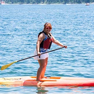 Person balancing on a paddleboard on clear blue water with pine trees in the background.