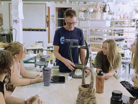 A group of people sit around a large table in a ceramics studio filled with pottery tools and shelves of clay pieces. One person at the center demonstrates a pottery technique using a clay piece, while others watch attentively. The table holds mugs, bottles, and sculpting tools.