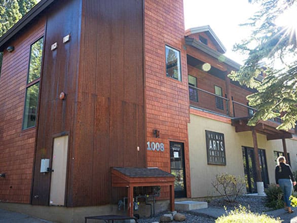 Exterior view of a two-story arts center building with a mix of wood and beige siding. The building has large windows, a balcony, and a sign reading “Holman Arts Center.” A person walks near the entrance, and sunlight filters through nearby trees.