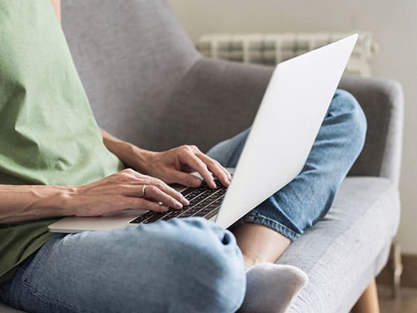 A person sits cross-legged on a cushioned chair, typing on a silver laptop. The individual is wearing a green sleeveless top and blue jeans. The background includes a radiator and part of a light-colored wall.