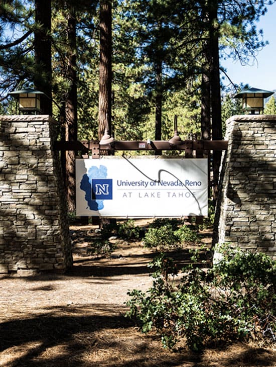 A stone entrance sign for the University of Nevada, Reno at Lake Tahoe campus. The sign is mounted between two stone pillars with lantern-style lights on top, surrounded by tall pine trees and natural landscaping.