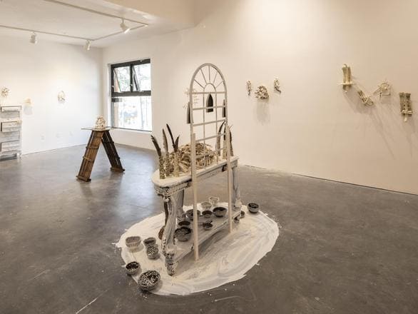 An art gallery space with white walls displaying sculptural pieces mounted in clusters. In the center, a decorative table with an arched frame holds mixed-media elements, surrounded by small ceramic bowls arranged on a circular white base. Additional artworks are placed on the walls and a wooden ladder-like structure stands near a window, creating an eclectic, contemporary exhibit.