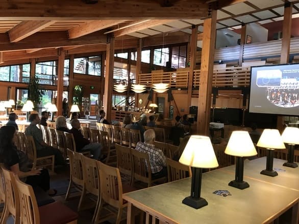 A group of people seated in wooden chairs inside the Prim Library at the University of Nevada, Reno, Lake Tahoe campus. The room has high wooden ceilings, large windows overlooking pine trees, and pendant lights. A projector screen displays a presentation at the front, while tables with lamps line the foreground, creating a cozy academic setting.