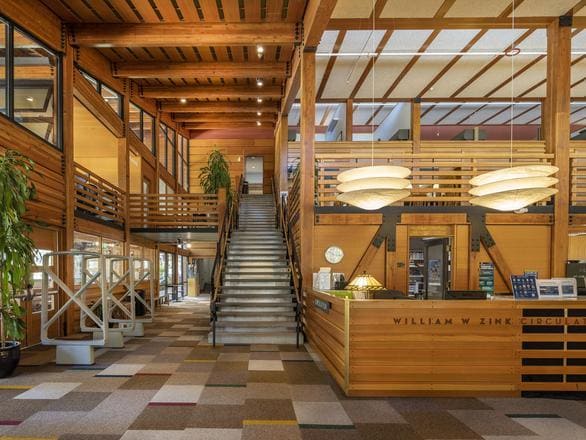 Interior of the Prim Library at the University of Nevada, Reno, Lake Tahoe campus. The space features warm wooden architecture with exposed beams, a central staircase leading to an upper level, and large windows allowing natural light. The William W. Zink circulation desk sits in the foreground, accented by modern pendant lights and colorful patterned carpet tiles.