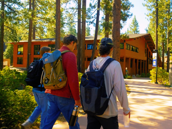 Students walking along a paved pathway on the Prim Library campus at the University of Nevada, Reno, Lake Tahoe. They carry backpacks and water bottles, surrounded by landscaped greenery and tall pine trees. In the background, a lodge-style building with wooden siding and large windows is visible under bright sunlight.