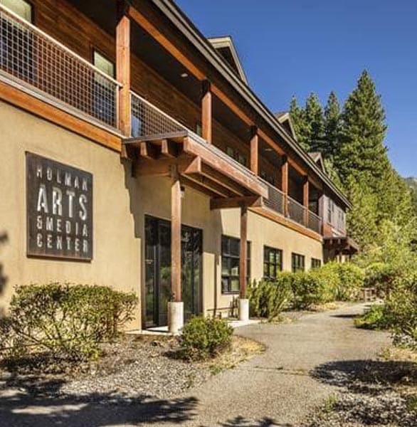 The Holman Arts & Media Center building at the University of Nevada, Reno, Lake Tahoe campus. The structure features a modern mountain-lodge design with beige stucco walls, warm wooden accents, and a second-story balcony framed by metal railings. A large sign reading “Holman Arts & Media Center” is mounted on the front wall. The building is surrounded by neatly trimmed shrubs and a gravel pathway, set against a backdrop of tall evergreen trees and a clear blue sky, evoking a serene alpine environment.
