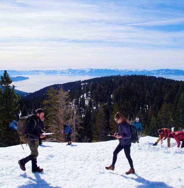 People conducting research on a snowy slope overlooking Lake Tahoe, with evergreen trees and distant mountains under a clear blue sky.