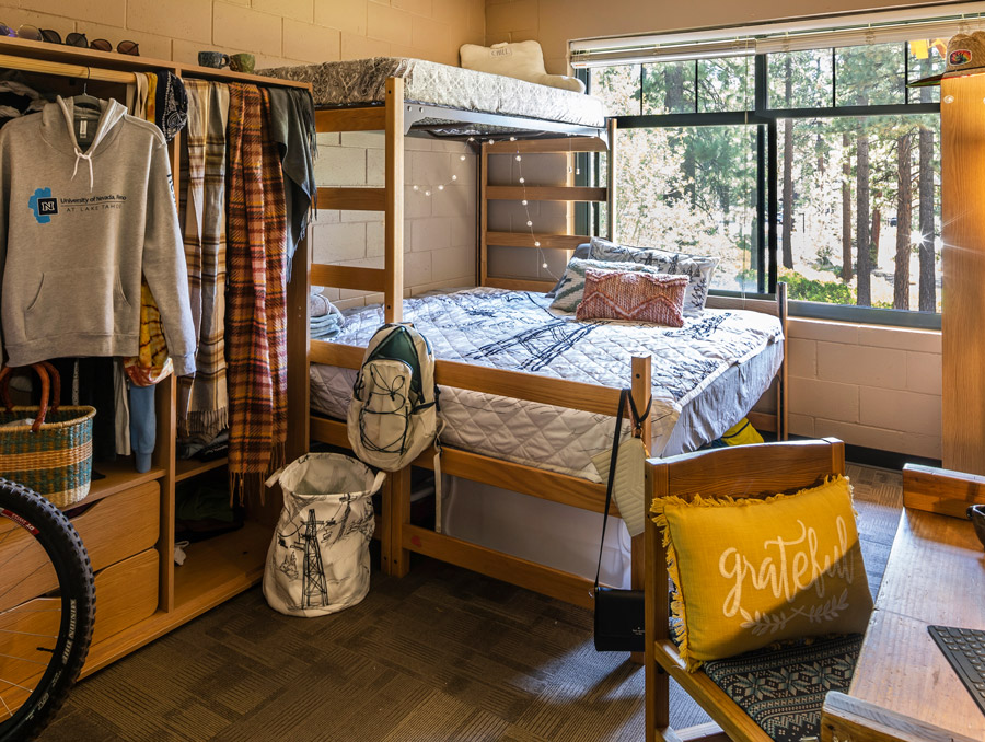 Prim-Schultz residence hall room with a wooden bunk bed, cozy bedding, and decorative pillows near a large window overlooking pine trees. Includes an open wardrobe with clothes and a hoodie, a desk with a chair featuring a “grateful” pillow, and personal items like a backpack and laundry bag.