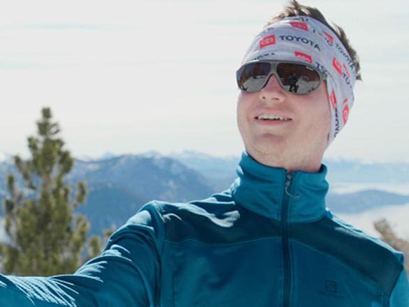 A person wearing a teal zip-up jacket and a white patterned headband standing outdoors in a snowy mountain setting. Evergreen trees are visible in the midground, and the background features distant snow-covered peaks under a bright, clear sky.