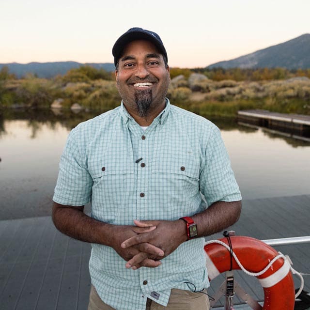 Sudeep Chandra standing on a wooden dock near calm water with a mountainous backdrop at sunset. The person is wearing a light green checkered short-sleeve shirt, tan pants, and a red wristwatch, with hands clasped in front. A bright orange life ring is attached to the dock railing on the right, and shrubs and trees line the shore in the background.