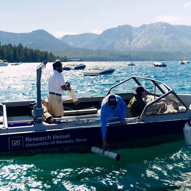 A University of Nevada, Reno research vessel floating on the clear blue waters of Lake Tahoe, with three people handling cylindrical sampling equipment. Several boats are anchored nearby, and forested mountains rise in the background under bright daylight.